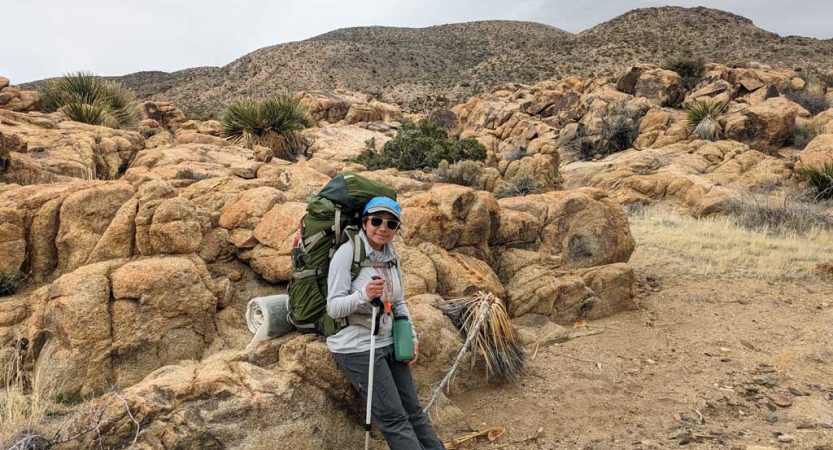 A person wearing a backpack rests against a large rock in the desert environment of Joshua Tree. 